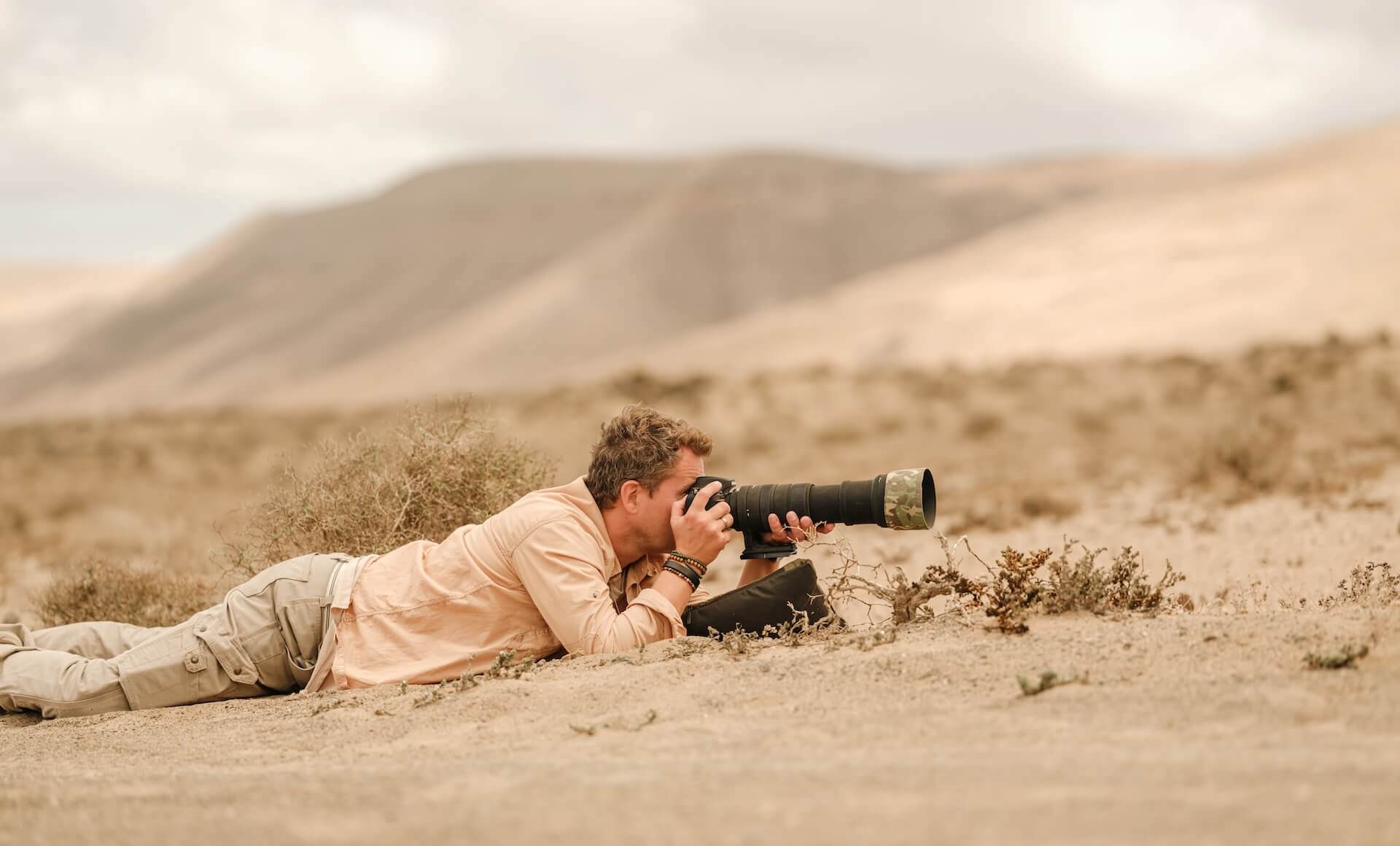 Persoon ligt op dorre grond te fotograferen met camera en telelens in een berglandschap.
