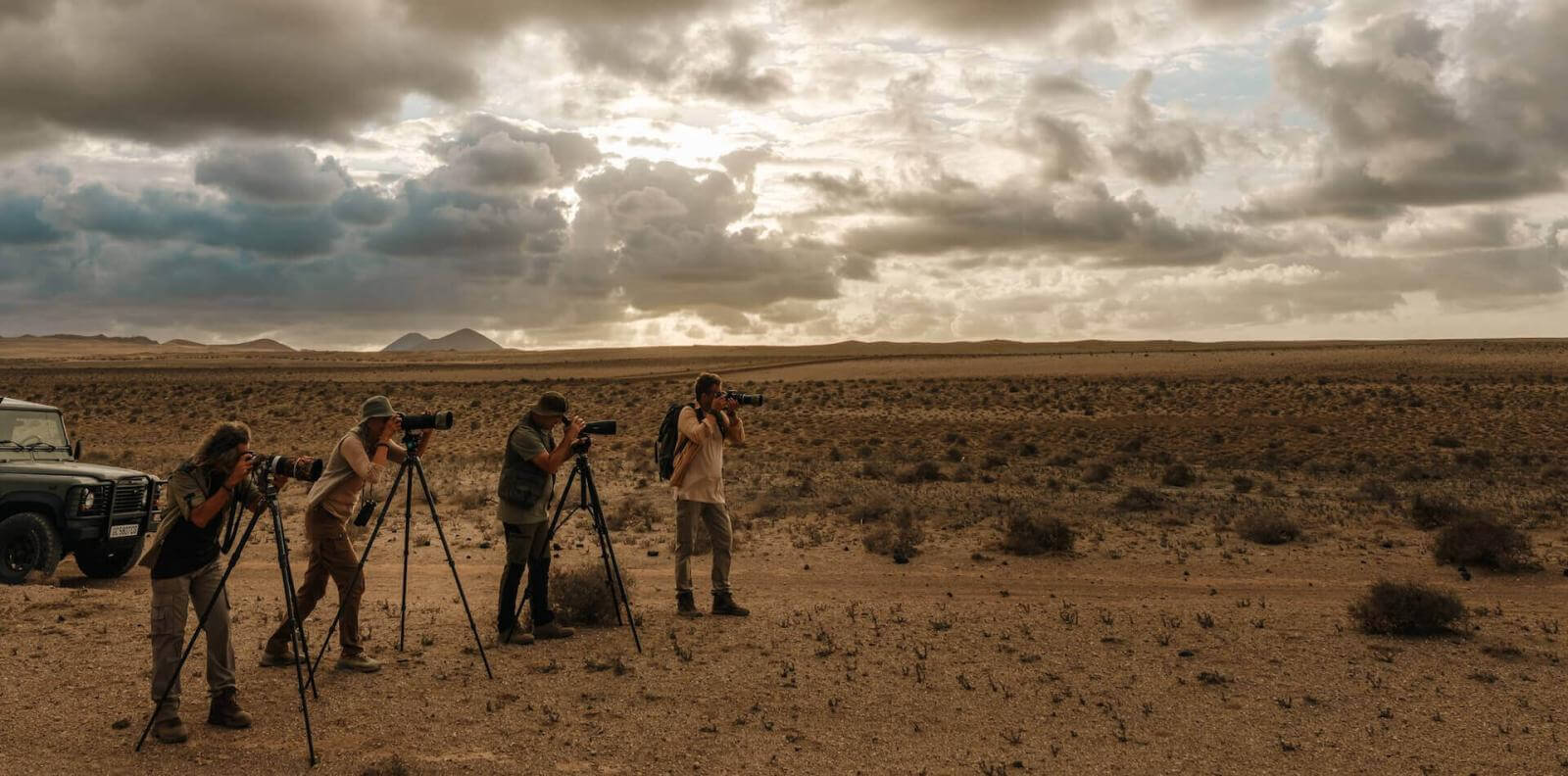 Vier mensen fotograferen vogels met telelenzen en statieven in een dor landschap bij zonsondergang.