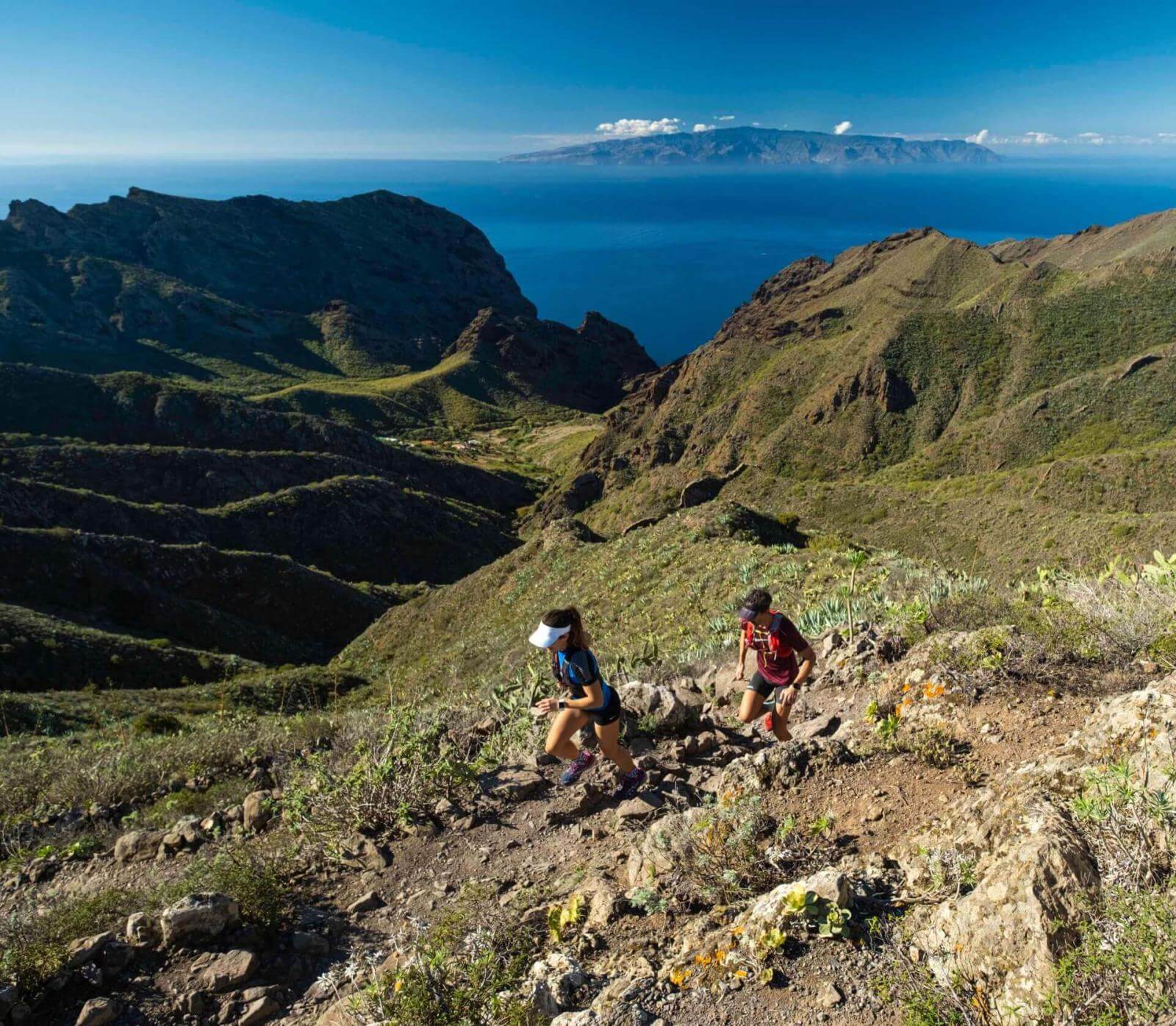 Twee mensen aan het trailrunnen op een bergpad met uitzicht op de oceaan en aan de horizon een ander eiland.