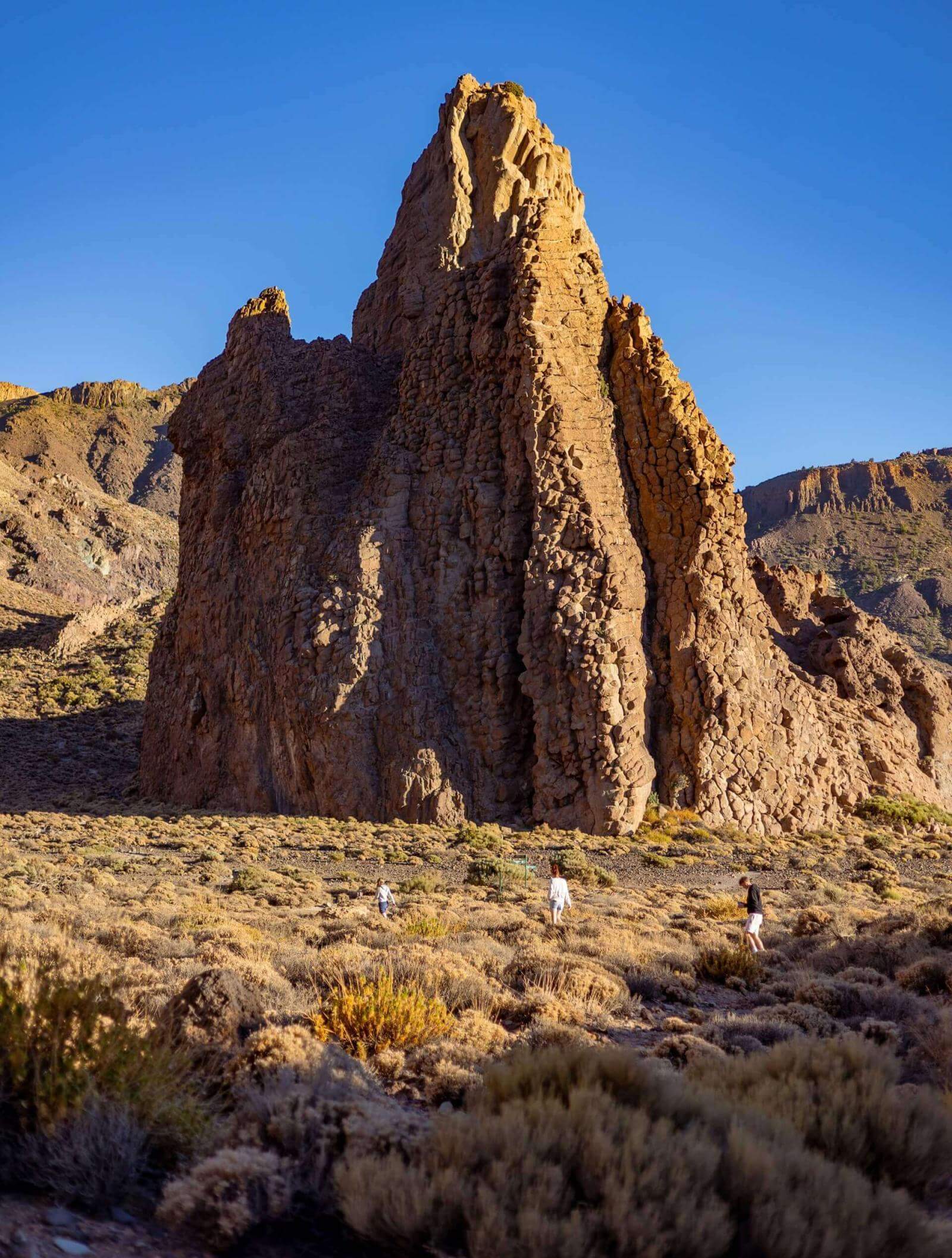 Mensen wandelen door laag struikgewas voor een grote verticale rotsformatie in een vulkanisch landschap.