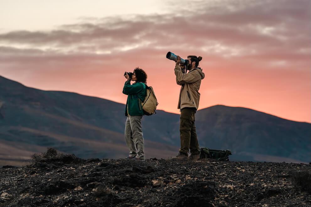 Twee mensen spotten vogels met een verrekijker en telelens in een vulkaanlandschap bij zonsopgang.
