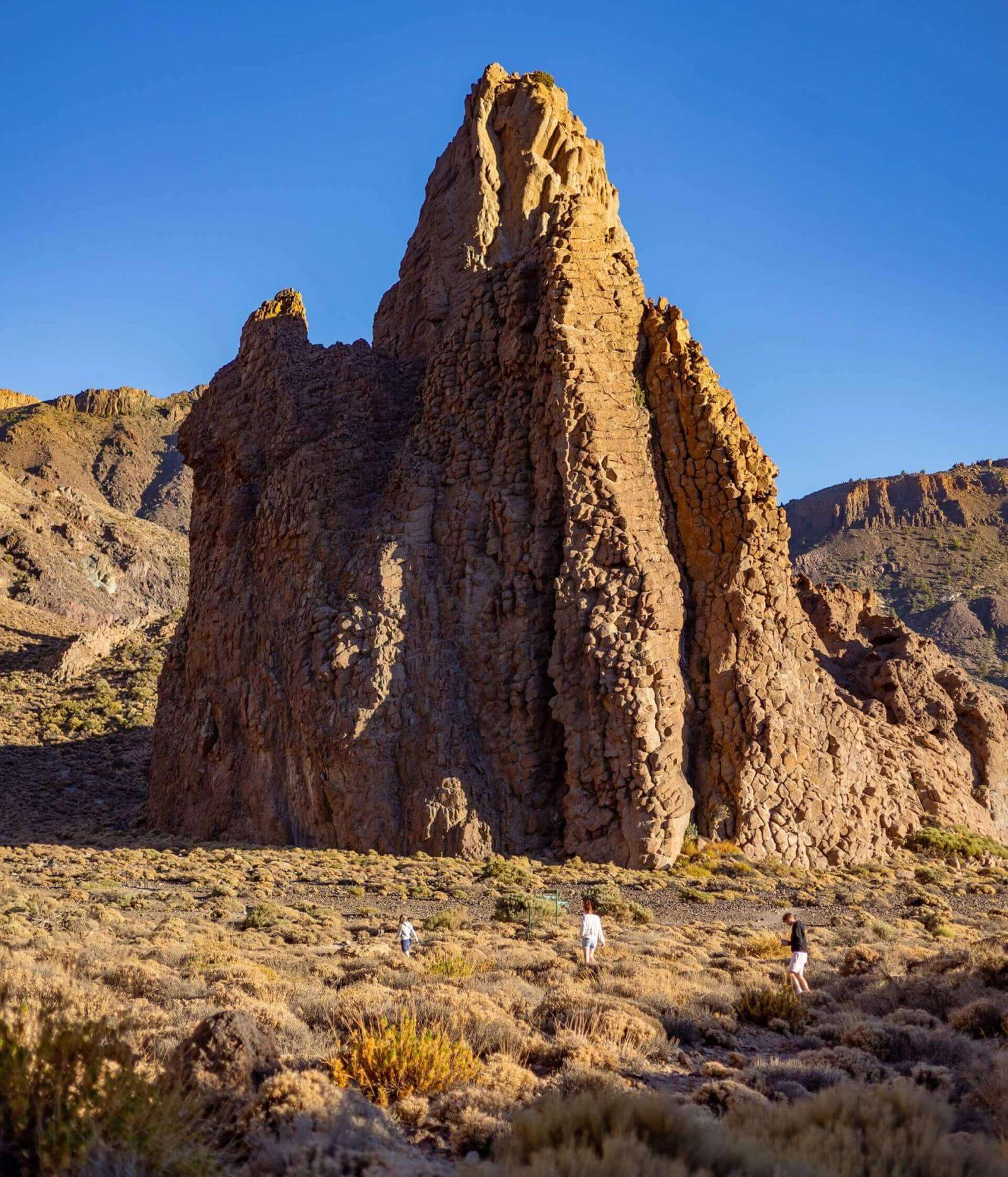 Mensen wandelen door laag struikgewas voor een grote verticale rotsformatie in een vulkanisch landschap.